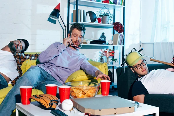 Young man with hangover adjusting eyeglasses while drunk friends sleeping on couches after home party — Stock Photo