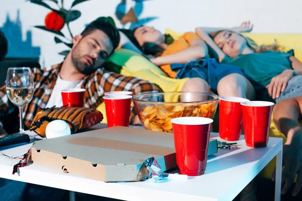Close-up view of pizza box, plastic cups and baseball equipment on table and drunk young people sleeping behind — Stock Photo