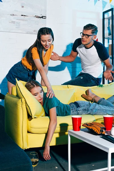 Smiling young woman waking up drunk girl sleeping on couch after home party — Stock Photo
