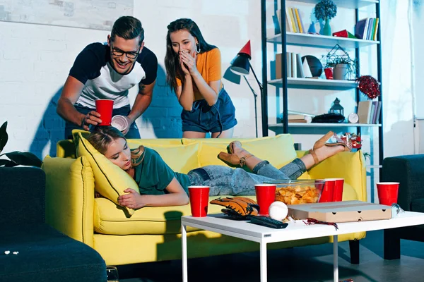 Lachende junge Leute, die betrunkene Freundin nach Heimparty auf Sofa schlafen sehen — Stockfoto