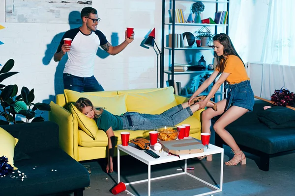 Smiling man holding plastic cups and looking at girl pulling drunk female friend sleeping on sofa after home party — Stock Photo