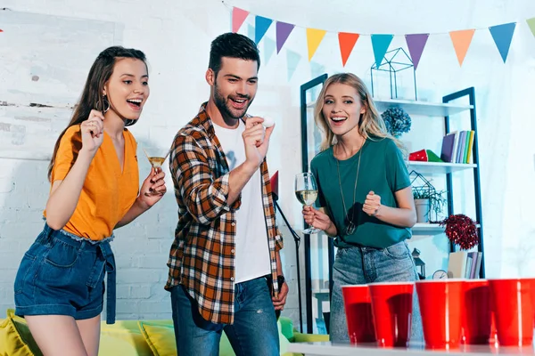 Усміхнені дівчата в келихах вина дивиться на молодого чоловіка, який грає пивний понг на домашній вечірці — стокове фото