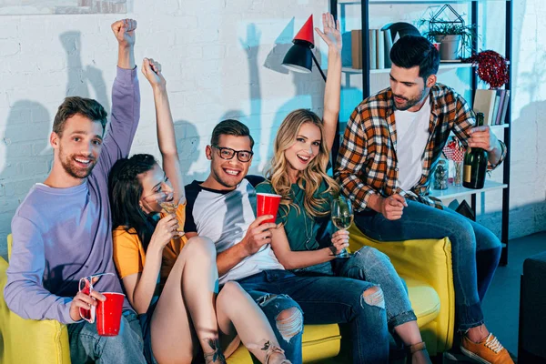 Heureux jeunes amis boire des boissons alcoolisées et sourire à la caméra à la fête à la maison — Stock Photo