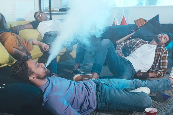 Young man smoking electronic cigarette while drunk friends sleeping after party — Stock Photo