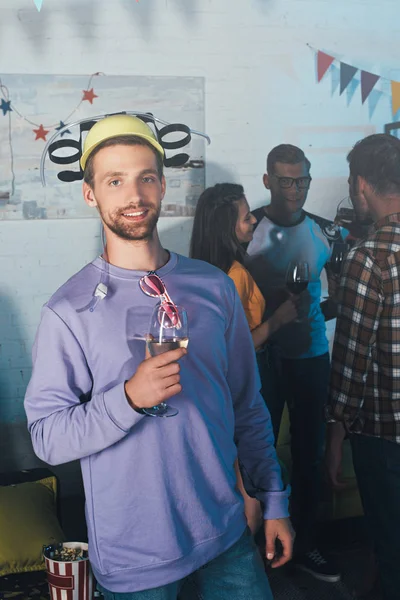 Jovem bonito segurando copo de vinho e sorrindo para a câmera na festa em casa — Fotografia de Stock