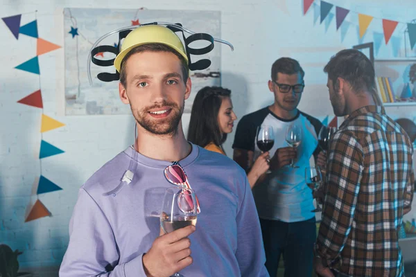 Joven guapo en sombrero de cerveza sosteniendo una copa de vino y sonriendo a la cámara en la fiesta en casa - foto de stock