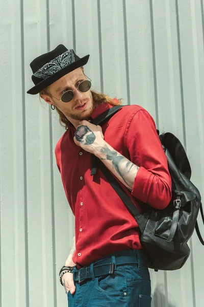 Stylischer tätowierter Mann mit Sonnenbrille und Hut mit Rucksack — Stockfoto