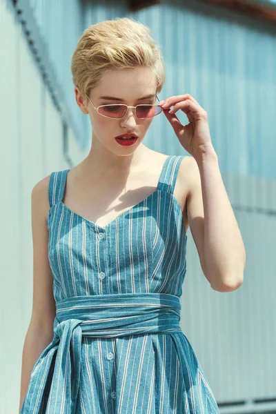 Fille élégante avec les cheveux courts posant en robe bleue à la mode et lunettes de soleil roses — Photo de stock