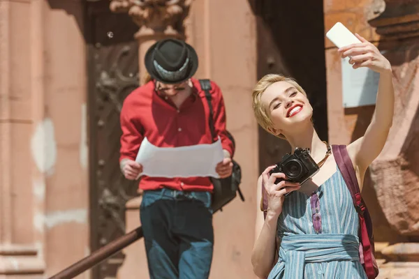 Девушка с камерой делает селфи на смартфоне, пока мужчина смотрит на карту — стоковое фото