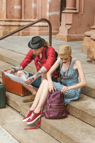 Couple of travelers with backpacks and vintage suitcases sitting on stairs and looking at map — Stock Photo