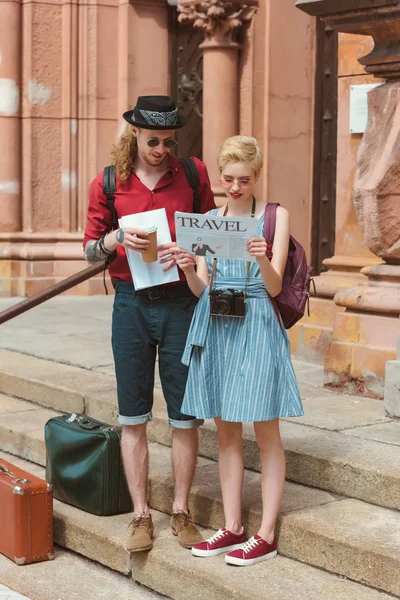 Un par de turistas con café leyendo el periódico de viaje mientras están de pie en las escaleras con maletas retro - foto de stock
