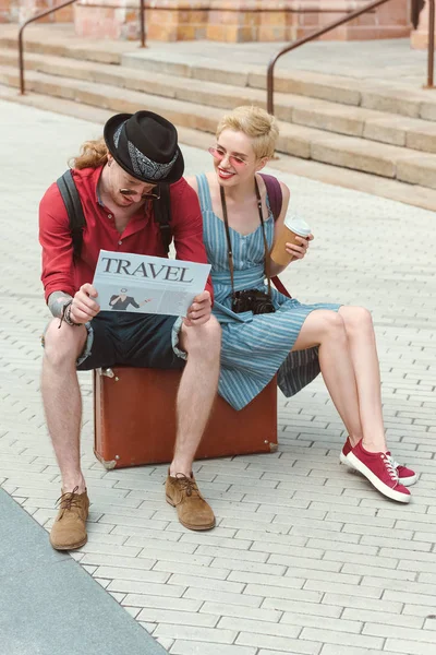 Couple of tourists with coffee to go reading travel newspaper and sitting on suitcase — Stock Photo