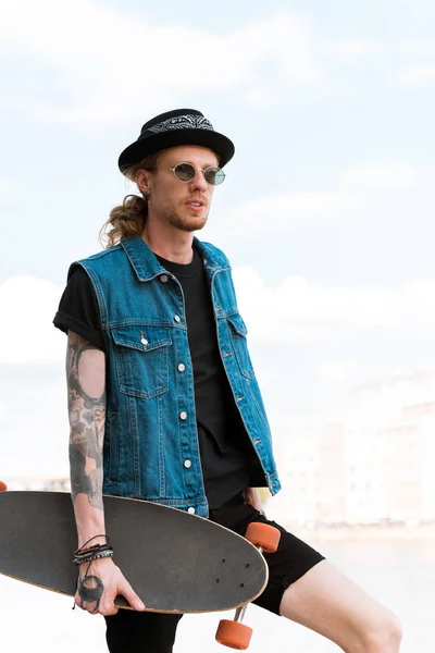 Bonito elegante tatuado homem de pé com skate e olhando para longe — Fotografia de Stock