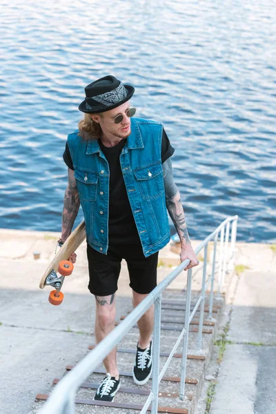 Handsome stylish tattooed man walking with skateboard on stairs near river — Stock Photo