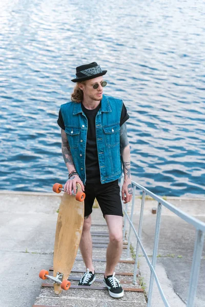 Handsome stylish tattooed man standing with skateboard on stairs near river — Stock Photo