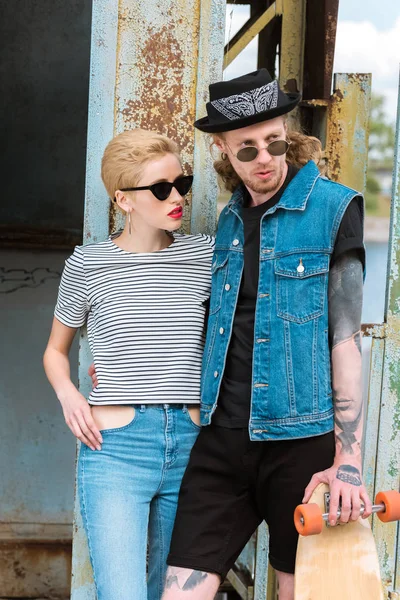 Novio con tatuajes y novia con estilo de pie con longboard y mirando hacia otro lado - foto de stock