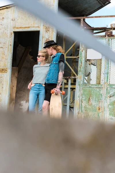 View through fence on boyfriend with tattoos and stylish girlfriend standing with longboard — Stock Photo