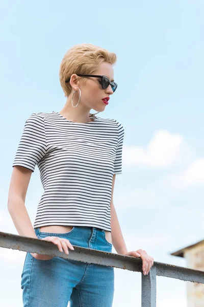 Chica con estilo en gafas de sol y con el pelo corto apoyado en barandilla - foto de stock