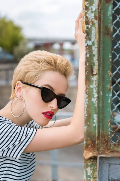 Stylish girl in sunglasses and with short hair leaning on fence — Stock Photo