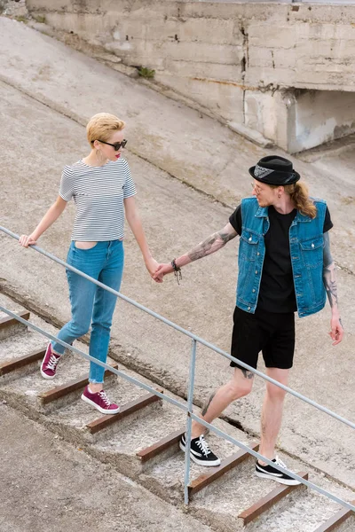 Freund mit Tätowierungen und stylische Freundin hält Händchen und geht Treppen — Stockfoto