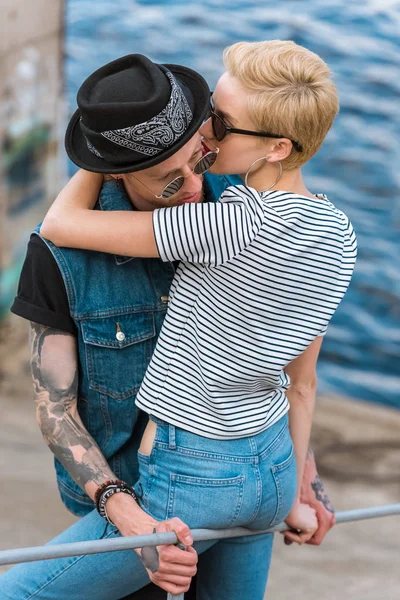 Boyfriend with tattoos and stylish girlfriend hugging and leaning on railing near river — Stock Photo