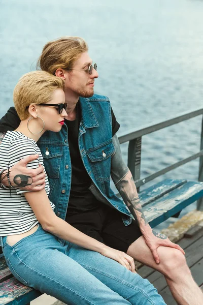 Vista lateral do namorado com tatuagens e elegante namorada abraçando no banco perto do rio — Fotografia de Stock