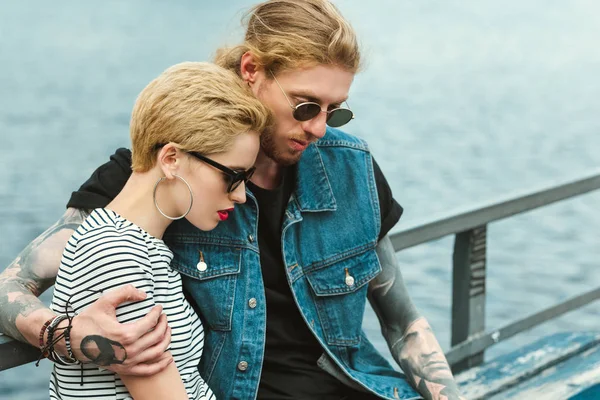 Boyfriend with tattoos and stylish girlfriend hugging near river and looking down — Stock Photo
