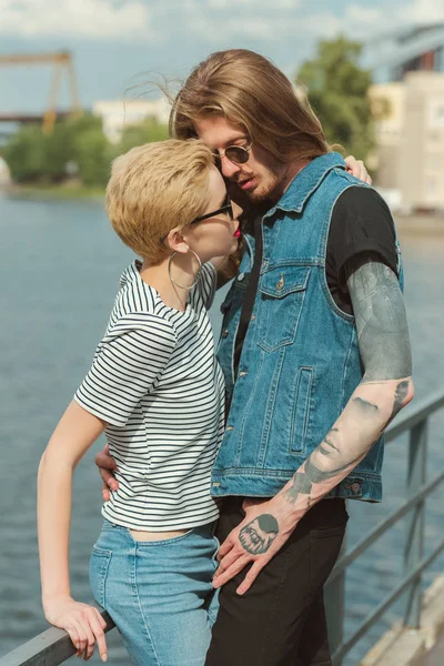 Copain avec des tatouages et copine élégante étreignant et allant embrasser sur le pont — Photo de stock