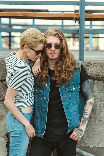 Novio con tatuajes y elegante novia posando en gafas de sol cerca del puente - foto de stock