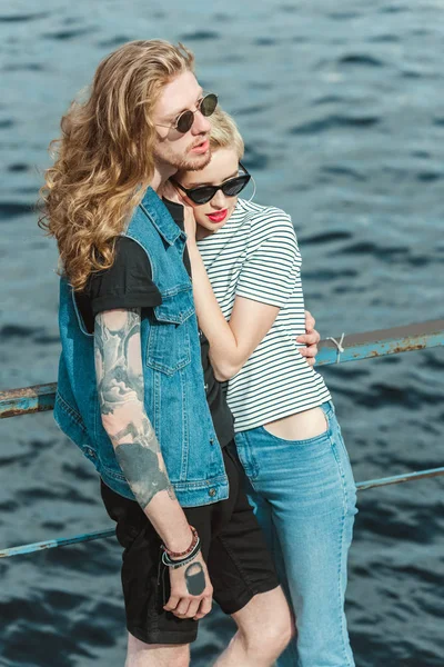 Vue grand angle de petit ami tatoué et copine élégante étreignant sur le pont près de la rivière — Photo de stock