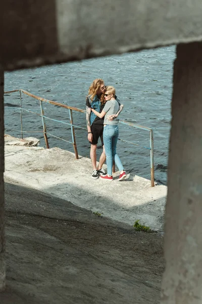 Copain tatoué et copine élégante étreignant sur le pont près de la rivière — Photo de stock