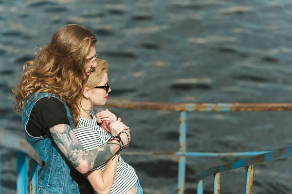 Vista laterale del fidanzato con tatuaggi che abbracciano ragazza elegante sul ponte vicino al fiume — Foto stock