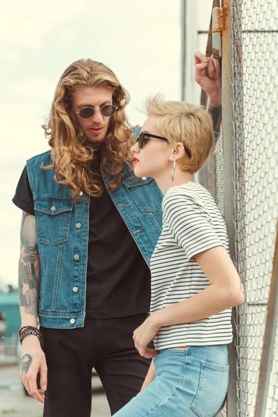 Novio con tatuajes apoyándose en la cerca y mirando a la novia con estilo - foto de stock