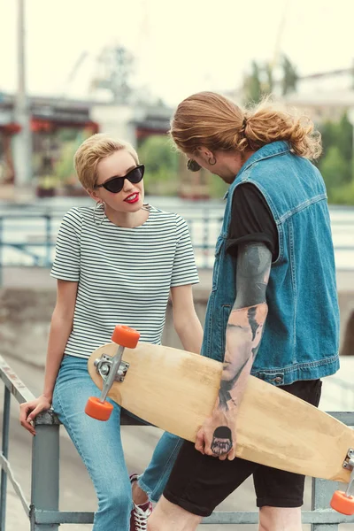 Petit ami avec tatouages tenant skateboard et regardant copine élégante — Photo de stock