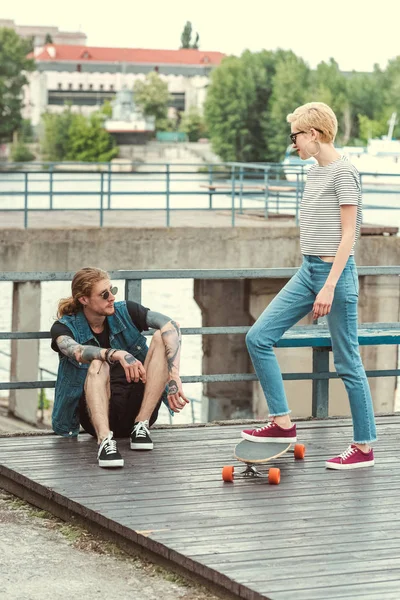 Elegante namorada colocando perna no skate e olhando para o namorado tatuado — Fotografia de Stock