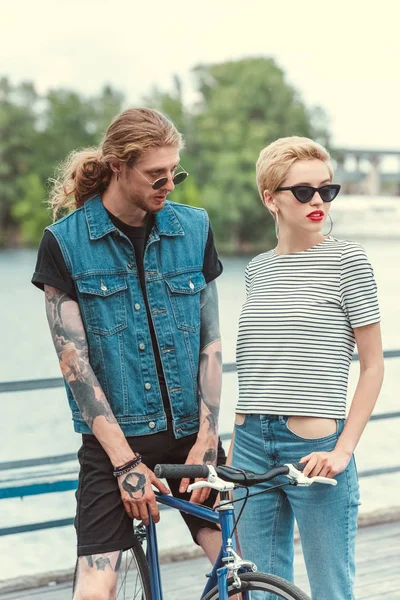 Novio con tatuajes y novia con estilo de pie con bicicleta en el puente y mirando hacia otro lado - foto de stock