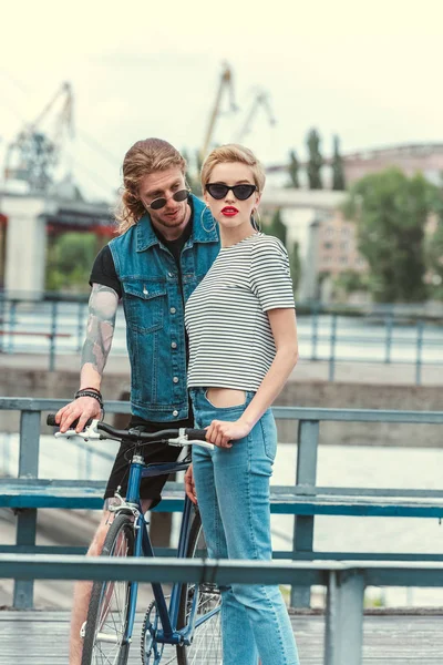 Novio con tatuajes y elegante novia posando cerca de la bicicleta en el puente - foto de stock