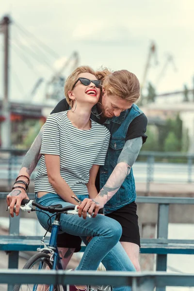 Copain tatoué et petite amie élégante souriante sur le vélo sur le pont — Photo de stock