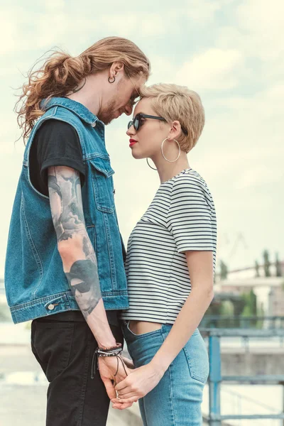 Low angle view of boyfriend with tattoos and stylish girlfriend holding hands and touching with foreheads — Stock Photo