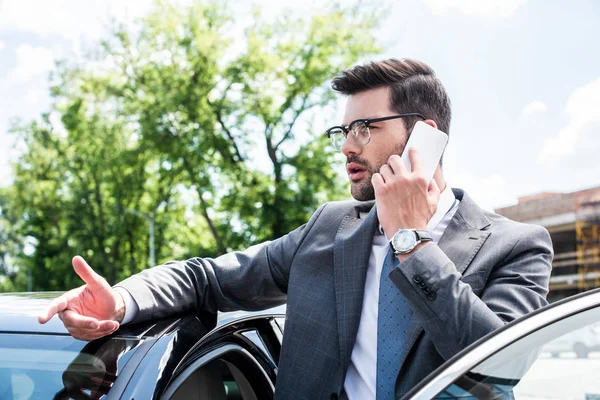 Uomo d'affari in occhiali parlare su smartphone mentre in piedi vicino auto sulla strada — Foto stock