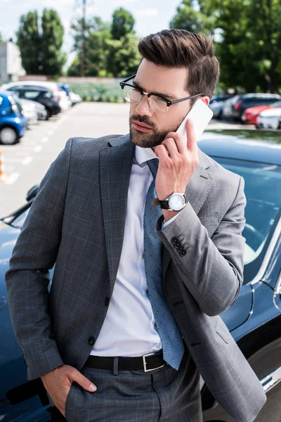 Portrait of businessman in eyeglasses talking on smartphone while standing near car on street — Stock Photo