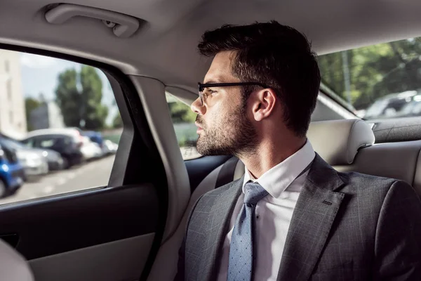 Ritratto di uomo d'affari pensieroso in occhiali che guarda fuori dal finestrino dell'auto mentre seduto sul sedile posteriore in auto — Foto stock