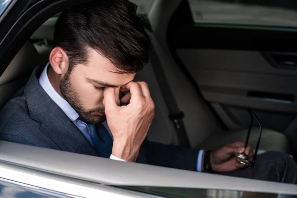 Side view of tired businessman with eyeglasses sitting on backseat of car — Stock Photo