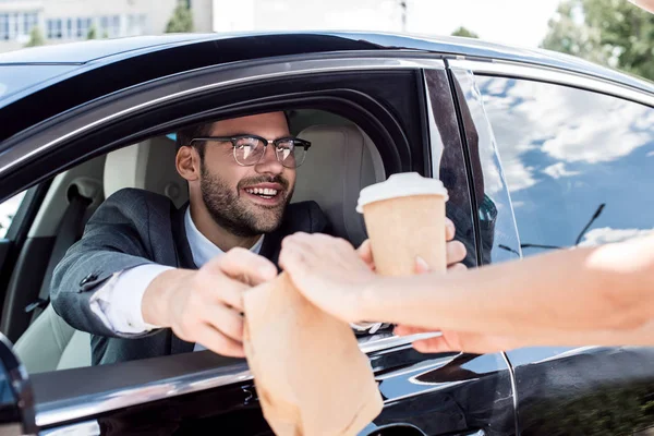 Обрізаний знімок усміхненого бізнесмена, який бере каву, щоб піти і забрати їжу від офіціанта, сидячи в машині — стокове фото