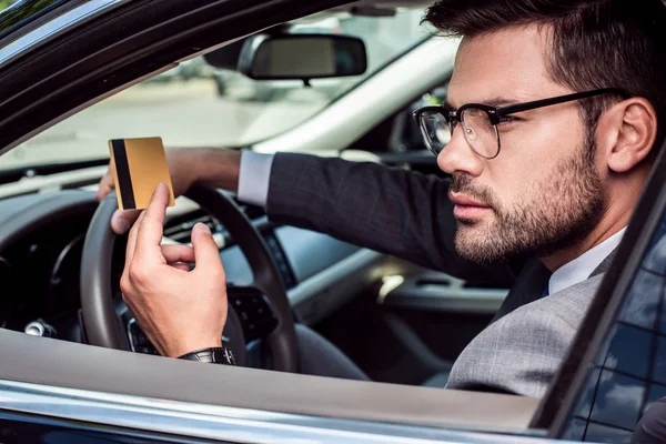 Vista lateral del hombre de negocios con tarjeta de crédito en la mano sentado al volante en el coche - foto de stock