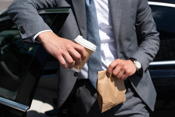 Vista parcial del hombre de negocios con comida para llevar y café para ir de pie en el coche - foto de stock