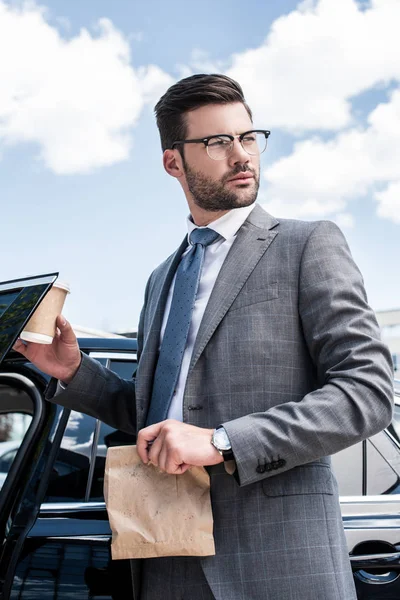 Businessman in eyeglasses with take away food and coffee to go standing near car on street — Stock Photo