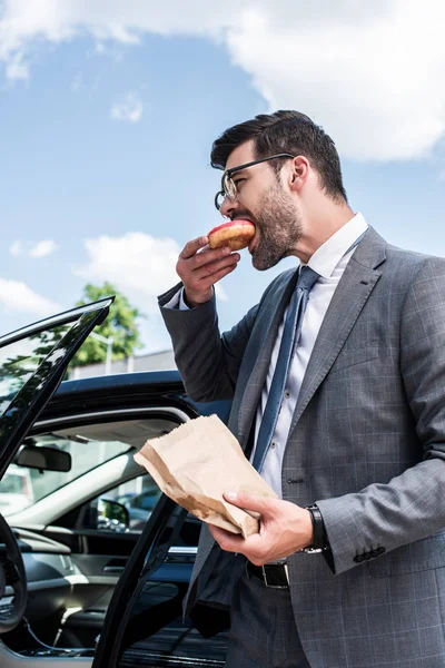 Seitenansicht eines Geschäftsmannes, der Donut zum Mitnehmen isst, während er auf der Straße am Auto steht — Stockfoto