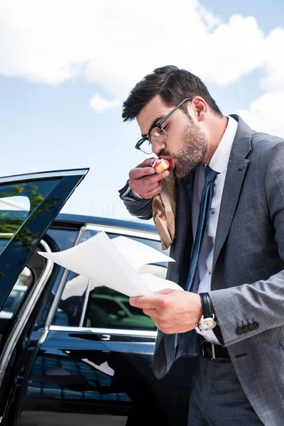 Geschäftsmann isst Donut und schaut sich Dokumente an, während er neben Auto auf der Straße steht — Stockfoto