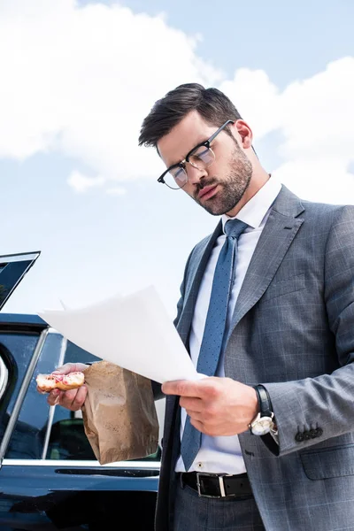 Businessman with doughnut looking at documents while standing near car on street — Stock Photo